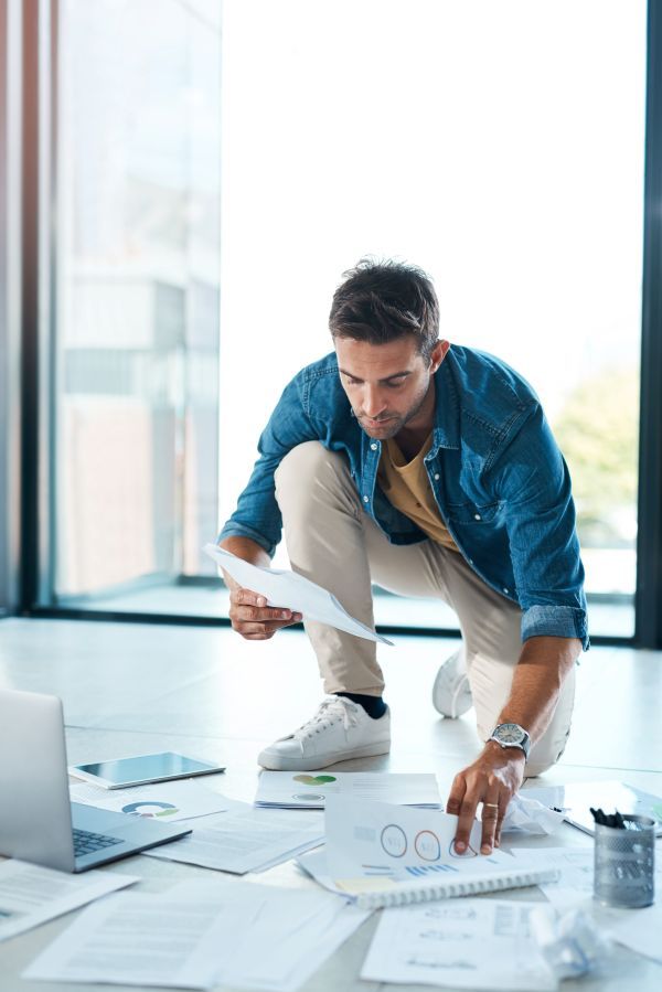 A man crouched on the floor with his laptop surrounded by paper